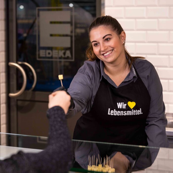 Auszubildende zur Frischespezialistin bei EDEKA Südwest steht hinter der Käsetheke und reicht einer Kundin ein Stück Käse